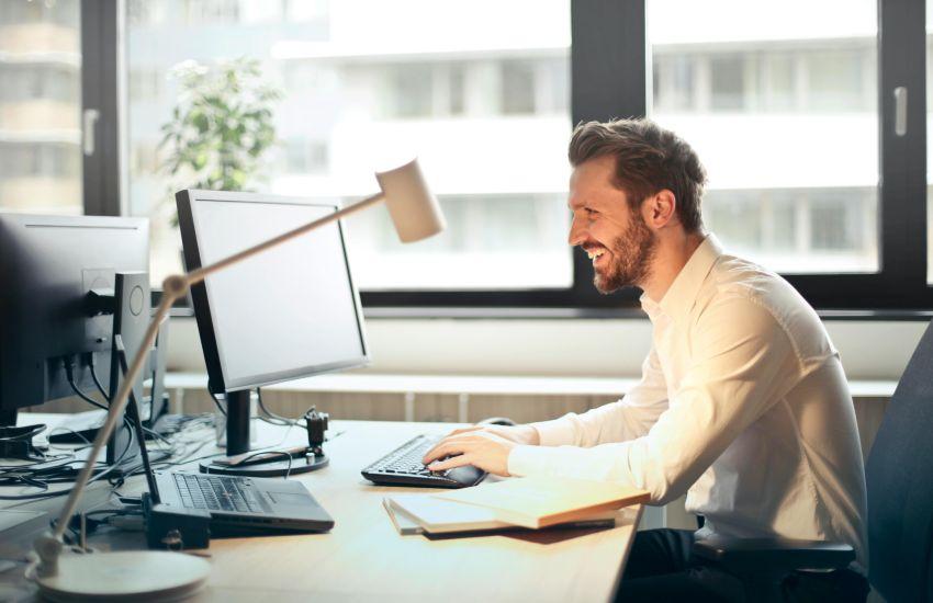 Man in White Dress Shirt Sitting on Black Rolling Chair While Facing Black Computer Set and Smiling.
